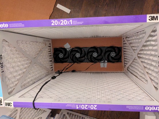 The view inside of the CR box from the (open) bottom. You can see a rectangular box made of 3m filtrete vent filters. The box is 20" long on one side, and 10" on the other side. At the bottom of the box (which, since it's upside-down, is actually the top) is the cardboard seen previously with a rectangle traced on it. Now there's a rectangle cut out of the cardboard, roughly following that traced line but maybe half an inch in. Thus the row of fans sits on the cardboard, with a 1/2" lip keeping…