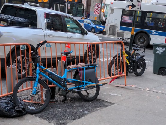 NYC corner. With cars/buses in the background, there's some ebikes parked to a sad pole while my bike (a blue Yuba mid-tail) is locked to an orange construction/crowd control barrier. Both the barrier and bike are a partially falling over.  The front wheel of the bike sits in a literal black bag of garbage.