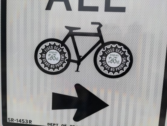 A (DOT and.. maybe MUTCD?) wayfinding sign for bikes; retro-reflective white with black text "ALL 🚲  →"

Over the wheels of the bicycle picture, someone has placed two round stickers that say, "FIXED GEAR.  NO GEARS NO PROBLEMS." and then something illegible "26st".