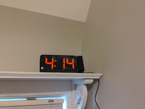A white shelf with a phone mounted on it. The phone, a generic black rectangle like every other smartphone, is sideways and is displaying the time '4:14' in red numbers.