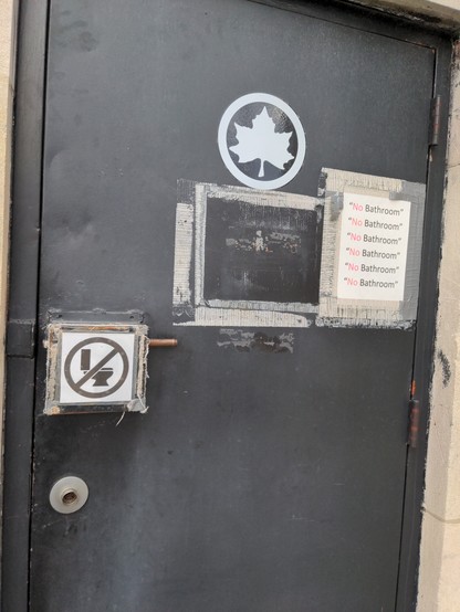 A black metal door with a NYC Parks logo (a maple leaf stencil) on it. On it there are two signs; one is a crossed-out toilet. The other says "No Bathroom" (with quotes), repeated 6 times.