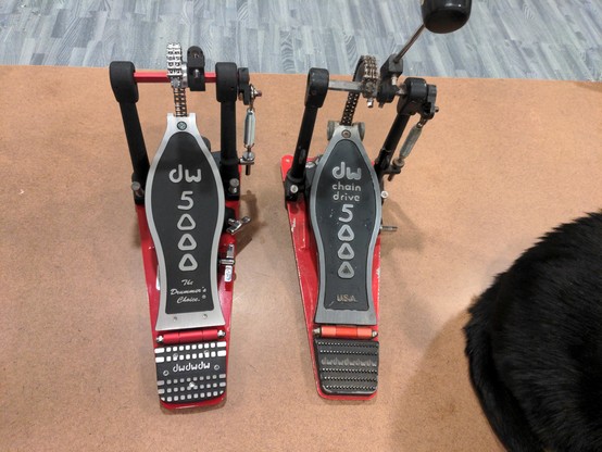 Two kick drum pedals, side by side (with a black cat butt corner photobomb). Both pedals have a red base, with the foot platform and components either painted black or shiny metal. The foot platform is attached to a chain, which then attaches to the beater that hits the drum head (although the one on the left doesn't have a beater installed yet).

The one on the left looks brand new, everything new and shiny. The pedal reads, "DW 5000 The Drummer's Choice". The one on the right looks old and wo…