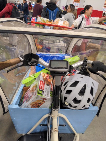 Bakfiet-style bike, filled with bulk groceries in the (blue) box; 2 trays of tofu, 30 pack of toilet paper, seaweed, etc. The view is from behind the box. A black/white/clear rain cover covers the box, and food is leaning against some of the sides of the rain cover. A white helmet hangs from the handlebars. In front of the bike, people lined up for the checkout register.