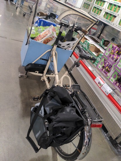 Same bike, but this time the shot from behind the rear wheel. A black pannier hangs from the rear rack. The bike frame is white, with a blue box on a black platform. A hailong battery sits atop the platform. Same groceries in the box.