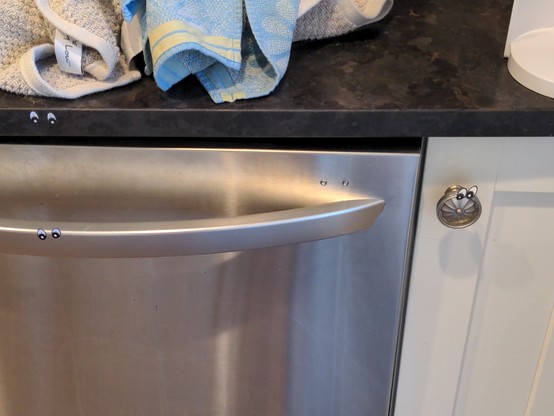 A black stone countertop with two eye stickers on the edge of it. Below that, a silver dishwasher handle with two eye stickers on it. To the right, a cabinet handle has, you guessed it, two eye stickers on it.