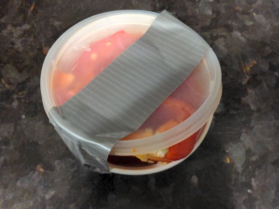 A plastic takeout container filled with something red. It's a white bottom with a clear lid; clearly different (and incompatible) pieces. Because it doesn't close properly, it is sloppily wrapped in gray duct tape.