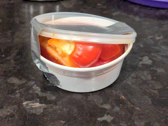 Side view of same container. You can see that it's overstuffed with red pepper; it should've been in a container twice the size. The tape is failing to keep the lid shut, and contents are spilling out.