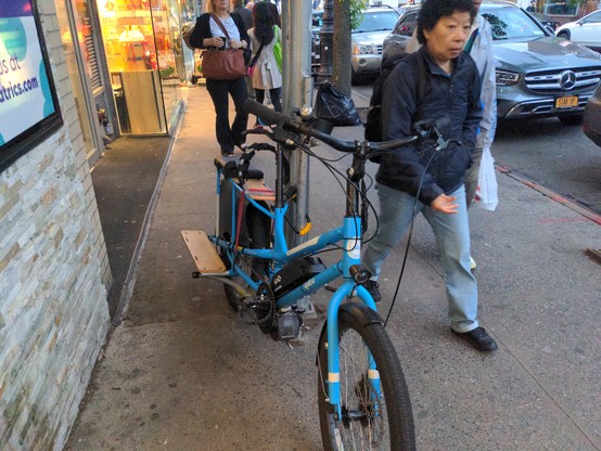 Blue mid tail bicycle (yuba kombi) locked to scaffolding on a sidewalk. The scaffolding divides the sidewalk by 1/3rd, and the bike blocks access to that side.

There are lots of people on the (too small) sidewalk, walking in both directions.