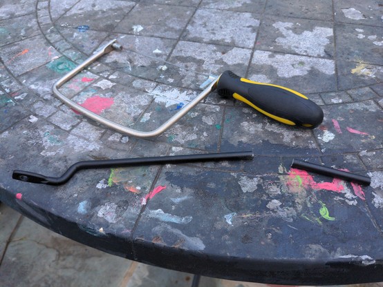 An outdoor patio table covered in coloful kids' paint, with a hacksaw and a black strut sitting on it. The strut has been cut about 2in from the end with the saw.