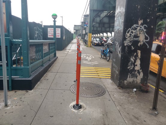 Sidewalk next to a roadway. On the left, a subway entrance. On the right, the roadway with large columns next to it (holding up an elevated subway). In the middle, the sidewalk is divided with delineator posts. The left side of the sidewalk is for marked for pedestrians, and the right side is marked for bikes (via sharrows, which is.. not what they fucking mean, NYC DOT assholes! Sharrows literally mean a shared lane BETWEEN CARS AND BIKES. The 2009 MUTCD states, "Shared Lane Markings shall not…