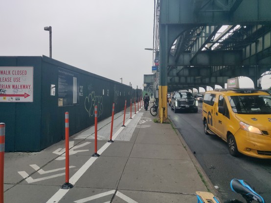 Further down the same sidewalk, viewed from the bike area. The left side no has a "sidewalk closed use pedestrian walkway" sign and some green walls, then the ped walkway, then the bike area, and then columns and then the roadway (with cars moving in it). The bikeway continues to narrow to 3-4ft wide, and there's a bike parked next to a column that further narrows the bikeway. Someone is biking just past it, to give you an idea of a 2.5ft wide cyclist just barely fitting (forget a cargo bike or…
