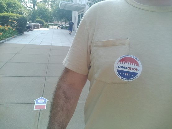 Person with a yellowish shirt on and an "I voted early!" sticker. Behind his arm, there's a sticker on the sidewalk with an arrow pointing to the polling station.