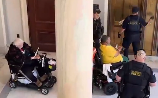 Screenshot from the video, zoomed in on two clearly elderly & disabled people sitting in power wheelchairs/mobility devices. They are surrounded by police in riot gear vests, and their hands are zip-tied together (despite being in, y'know, wheelchairs).