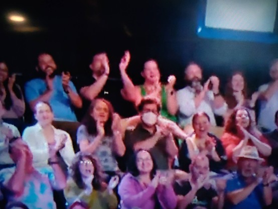 An audience clapping at a Daily Show taping, zoomed into the top 3 rows. In the middle, a guy wears a white N94 mask (surrounded by maskless people).