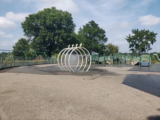 A NYC playground in full sun (there are kids there , but out of frame in the shade). There are 4 metal rings 7 ft tall spraying water for kids to run through. But the way they're aligned, it almost looks like a tiny car wash.