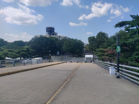 View from an overpass bridge sidewalk, showing a chain-link fence at the end completely blocking access to the park. In the background, trees and the big.. I don' t even know what they are, sculptures?