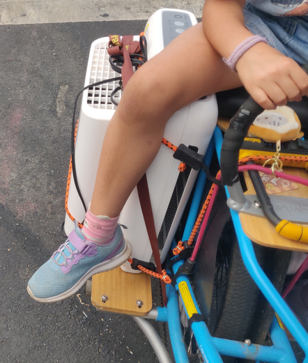 The rear of a blue Kombi mid-tail cargo bike. Along the rear right side boards at the bottom, a white air filter unit (Coway 200M) is bungeed to the side of the bike. The bottom half of a kid is also visible, sitting on the rear rack, her leg over the air filter, a hand on the ring to hold on.